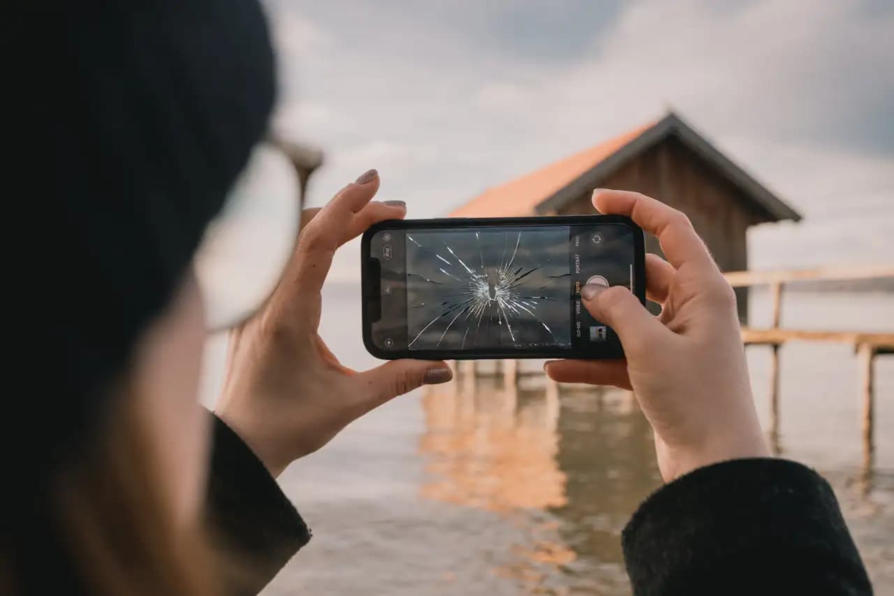 Osoba robiąca zdjęcie za pomocą telefonu z pękniętą szybą na ekranie, w tle widok jeziora i drewnianego domku.
