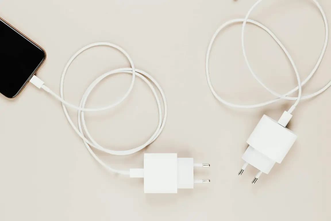 Białe ładowarki sieciowe z przewodem USB-C — jedna podłączona do smartfona, druga leży obok na jasnym tle.