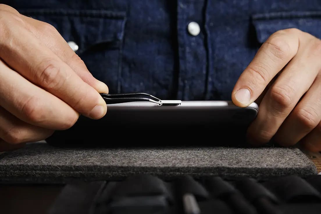Osoba w jeansowej koszuli używa metalowego narzędzia do otwarcia telefonu podczas samodzielnej naprawy.