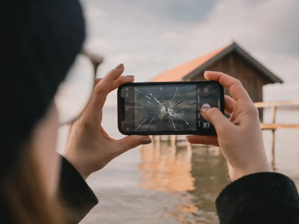 Osoba robiąca zdjęcie za pomocą telefonu z pękniętą szybą na ekranie, w tle widok jeziora i drewnianego domku.