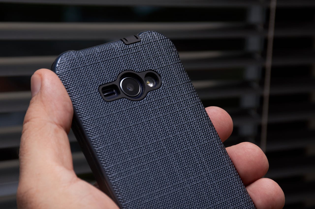 Smartfon w ochronnym teksturowanym etui z widocznym obiektywem aparatu i lampą błyskową, trzymany w dłoni. Czarne etui z charakterystycznym wzorem chroni telefon przed uszkodzeniami dzięki podwyższonym krawędziom wokół modułu aparatu.