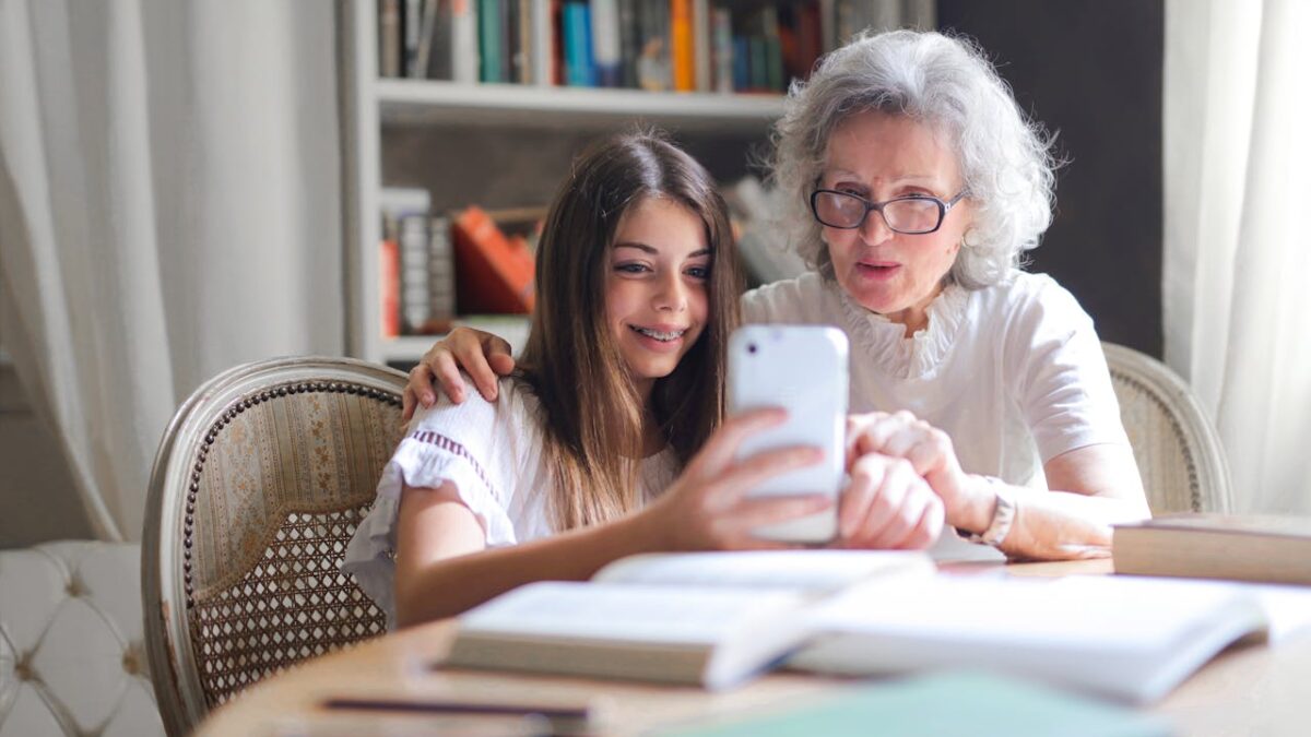 Młoda dziewczyna pomaga seniorce w obsłudze iPhone'a, siedząc razem przy stole w przytulnym pokoju z biblioteczką w tle. Obie uśmiechnięte patrzą na ekran telefonu podczas wspólnej nauki.