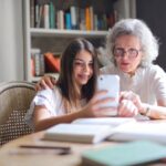 Młoda dziewczyna pomaga seniorce w obsłudze iPhone'a, siedząc razem przy stole w przytulnym pokoju z biblioteczką w tle. Obie uśmiechnięte patrzą na ekran telefonu podczas wspólnej nauki.