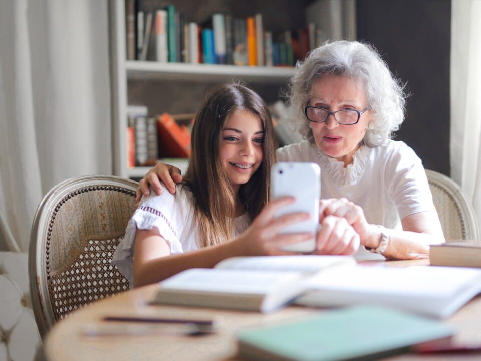 Młoda dziewczyna pomaga seniorce w obsłudze iPhone'a, siedząc razem przy stole w przytulnym pokoju z biblioteczką w tle. Obie uśmiechnięte patrzą na ekran telefonu podczas wspólnej nauki.
