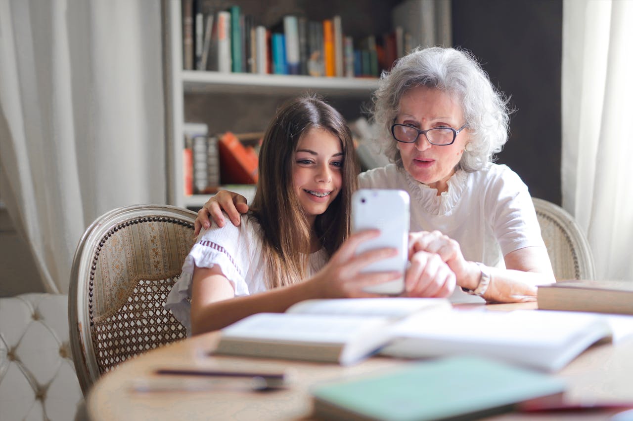 Młoda dziewczyna pomaga seniorce w obsłudze iPhone'a, siedząc razem przy stole w przytulnym pokoju z biblioteczką w tle. Obie uśmiechnięte patrzą na ekran telefonu podczas wspólnej nauki.