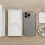 Używany iPhone 13 Pro w kolorze grafitowym z kompletnym zestawem: oryginalne pudełko, wkładka, kabel USB-C i dokumentacja na drewnianym blacie.