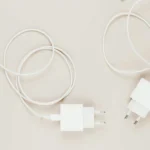 Białe ładowarki sieciowe z przewodem USB-C — jedna podłączona do smartfona, druga leży obok na jasnym tle.