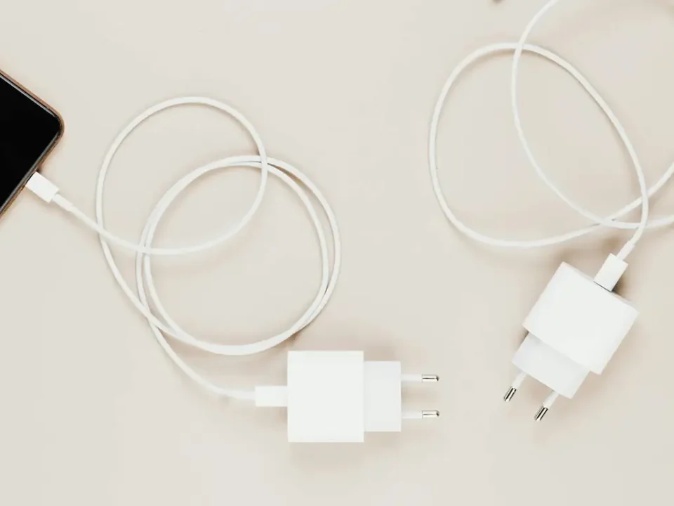 Białe ładowarki sieciowe z przewodem USB-C — jedna podłączona do smartfona, druga leży obok na jasnym tle.