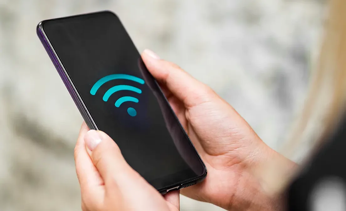 Osoba trzyma w dłoniach smartfon z wyświetlonym niebieskim symbolem WiFi na czarnym ekranie.