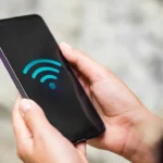 Osoba trzyma w dłoniach smartfon z wyświetlonym niebieskim symbolem WiFi na czarnym ekranie.