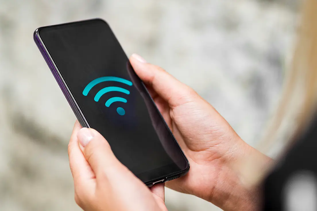 Osoba trzyma w dłoniach smartfon z wyświetlonym niebieskim symbolem WiFi na czarnym ekranie.