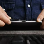Osoba w jeansowej koszuli używa metalowego narzędzia do otwarcia telefonu podczas samodzielnej naprawy.