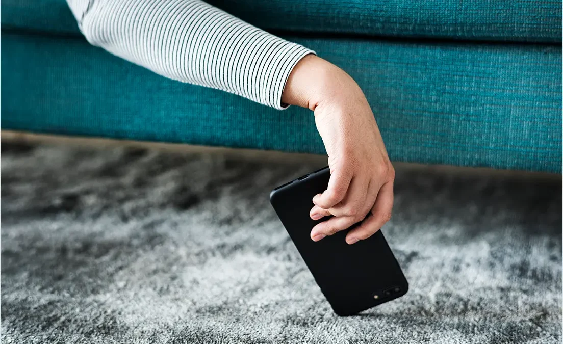 Osoba siedząca na turkusowej sofie podnosi z szarego dywanu czarny smartfon, który spadł na podłogę.
