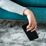 Osoba siedząca na turkusowej sofie podnosi z szarego dywanu czarny smartfon, który spadł na podłogę.