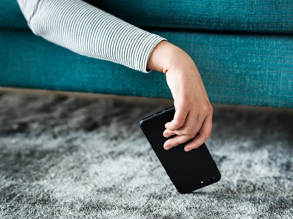 Osoba siedząca na turkusowej sofie podnosi z szarego dywanu czarny smartfon, który spadł na podłogę.