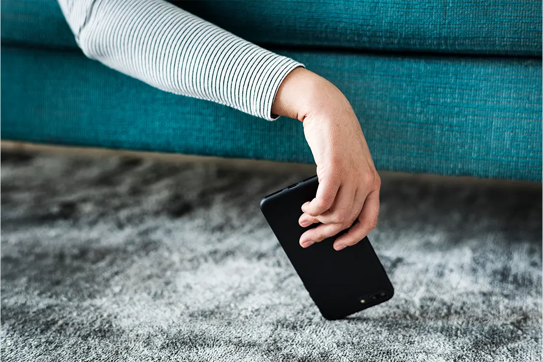 Osoba siedząca na turkusowej sofie podnosi z szarego dywanu czarny smartfon, który spadł na podłogę.